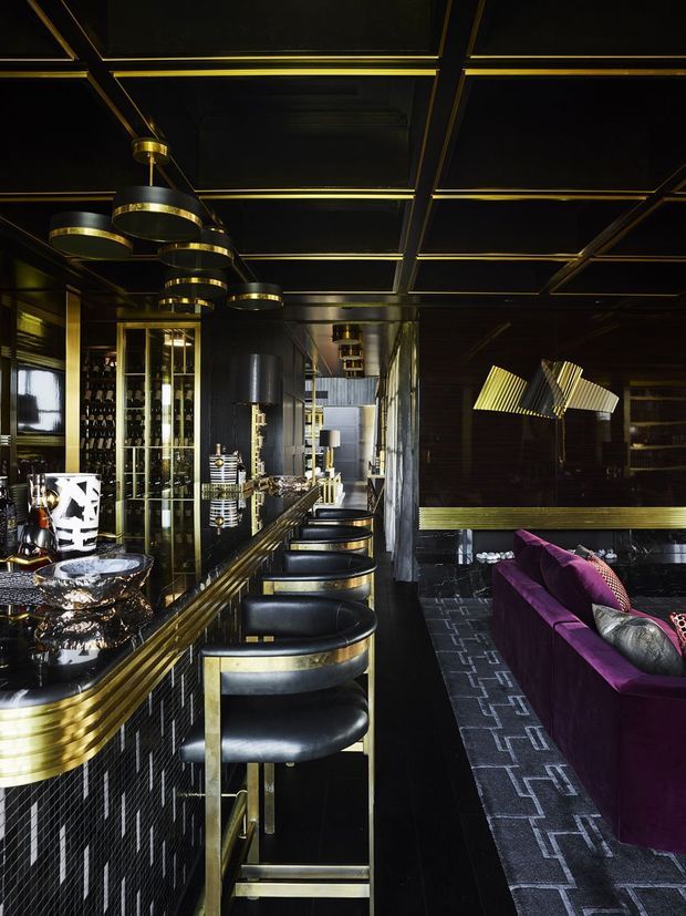A dramatic, richly detailed home bar and lounge with a long black-and-gold bar, upholstered bar stools, mirrored and glass-fronted bottle storage, and dark geometric wall and ceiling detailing. A deep purple sofa and sculptural gold lighting define the adjacent seating area. The overall atmosphere is luxurious, theatrical, and art-deco inspired.