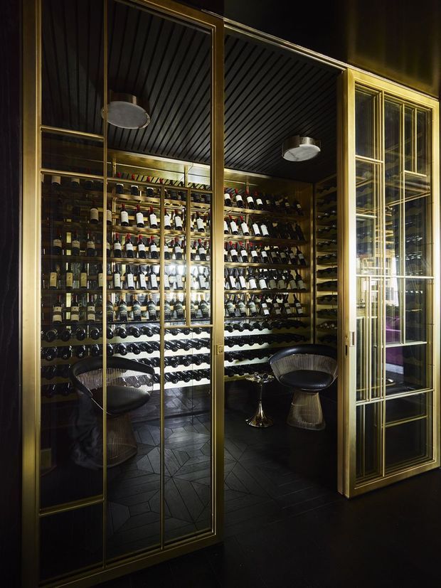 A dark, luxurious wine storage room enclosed by tall glass-and-gold framed doors. The room is densely lined with wine bottles on built-in shelving, with two sculptural dark lounge chairs, a black geometric tiled floor, and a dark ribbed ceiling. Warm integrated lighting highlights the metallic frames and wine collection.