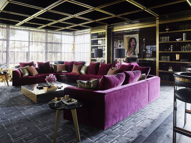 A dramatic, luxurious living room with deep plum velvet sectional sofas arranged around low coffee tables. The room features extensive dark built-in shelving, metallic trim details, floor-to-ceiling sheer glazing, a dark patterned carpet, and a striking geometric ceiling grid with gold-toned accents. A bar-height seating area is visible at the right.