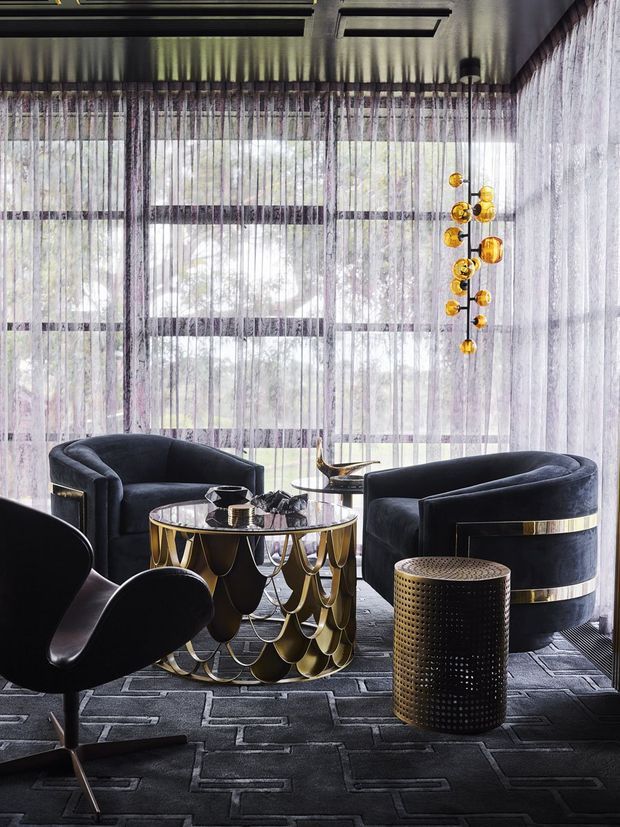 A luxurious, moody sitting area with sculptural black velvet lounge chairs and sofas arranged around a highly decorative round coffee table with gold metallic detailing. Floor-to-ceiling sheer curtains cover expansive glazing, while a clustered amber glass pendant hangs beside the seating. The room features a dark patterned carpet, a reflective dark ceiling with recessed linear lighting, and strong black-and-gold Art Deco-inspired detailing.