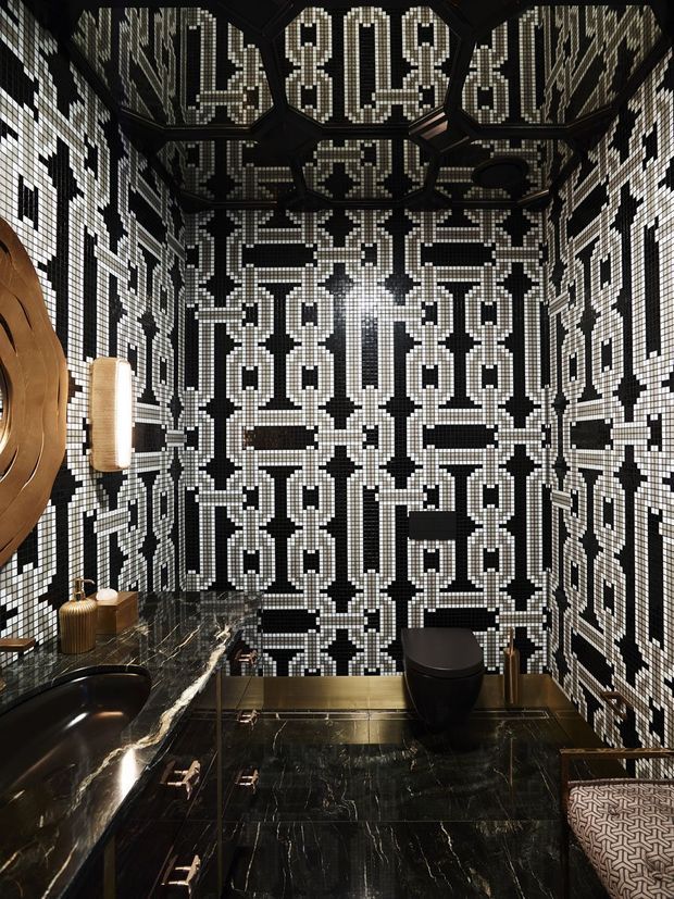 A dramatic, compact bathroom clad in an intricate black-and-white geometric mosaic tile pattern across the walls and ceiling. The room features a black marble vanity countertop with an integrated dark basin, dark cabinetry, a black toilet, a warm illuminated wall sconce, reflective dark marble flooring, and a small upholstered stool. The design is highly graphic, luxurious, and high-contrast.