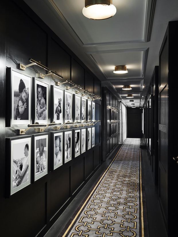 A long, narrow, dramatically lit interior hallway with dark paneled walls, ornate coffered ceiling detailing, repeated flush-mount ceiling lights, and a patterned runner carpet. One wall is arranged as a gallery with numerous black-and-white framed photographs illuminated by individual picture lights, while the opposite side contains dark paneled doors or cabinetry.