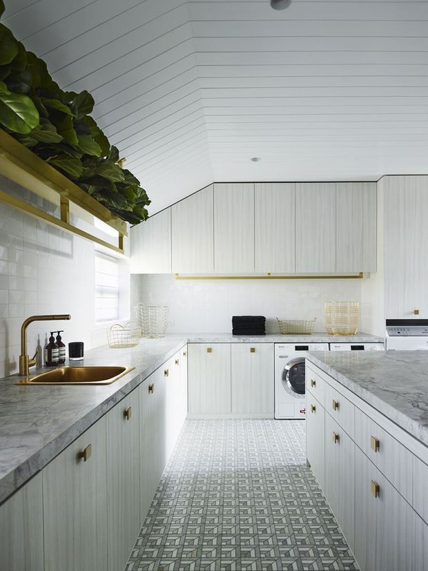 Bright, compact contemporary kitchen-laundry with pale vertically grained cabinetry, white tiled splashback, grey marble-look benchtops, brass hardware and tapware, patterned geometric floor tiles, and a white slatted ceiling. A washing machine is integrated along the rear cabinetry, while greenery sits on an upper shelf near the window.