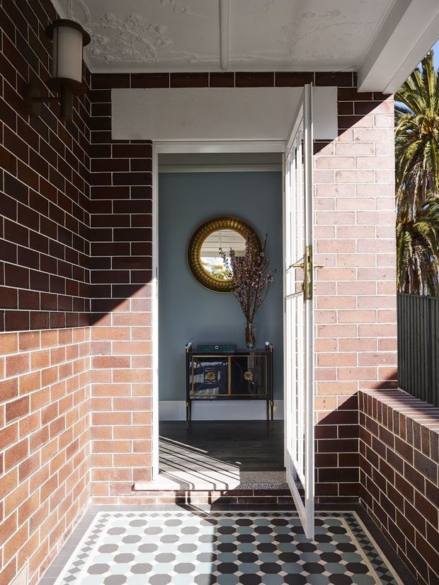 A sunlit residential entryway viewed from the exterior, framed by dark red and pale brick walls. The porch has patterned geometric floor tiles, a white decorative pressed-metal ceiling, and an open glazed door leading to a blue-grey hallway. Inside is a dark decorative console with a round gold-framed mirror and vase of branches. Palm foliage is visible beyond the side boundary.