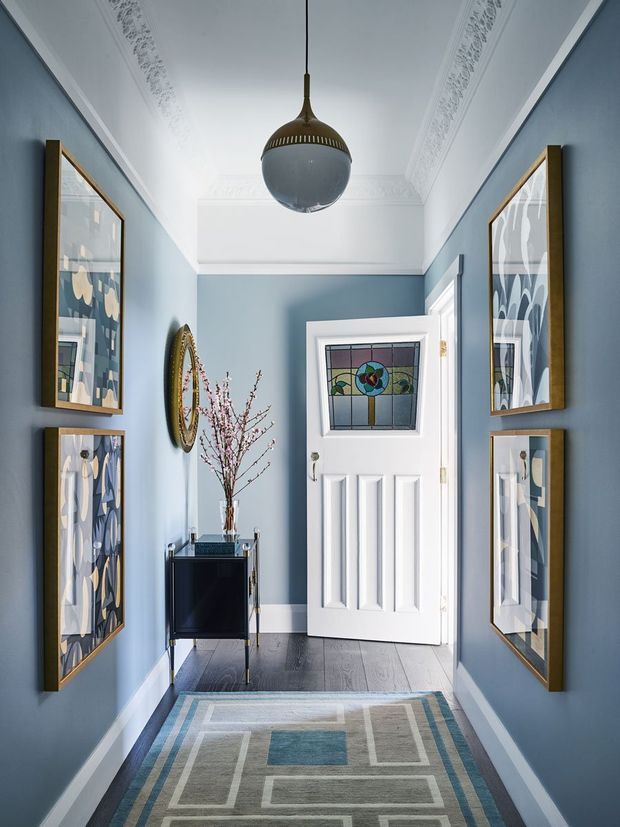 A narrow, elegantly styled hallway with dusty blue walls, white ornate ceiling cornices, dark timber flooring, and a white paneled entry door featuring a stained-glass window. Framed abstract artwork lines both walls, while a small dark console cabinet, oval mirror, decorative branches, pendant light, and geometric runner add refined character.