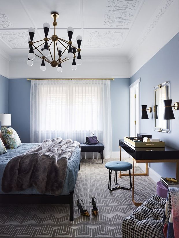 A refined bedroom with pale blue walls, ornate white decorative ceiling panels, and a dramatic brass-and-black geometric chandelier. The room features a bed with a blue upholstered cover and layered grey throws, a patterned grey rug, sheer curtains, a dark console desk or dressing table, wall sconces, a small upholstered stool, and a patterned armchair. The overall look is elegant, tailored, and slightly Art Deco-inspired.