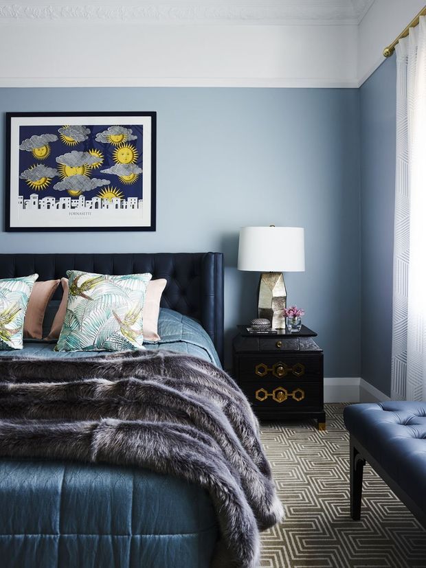 A stylish bedroom with deep blue walls, a tufted navy upholstered bed, layered botanical cushions, and a dark faux-fur throw. A black and gold bedside table with a sculptural lamp sits beside the bed, while a matching tufted bench occupies the foreground. Framed artwork, white sheer curtains with a brass rod, decorative ceiling cornice, and a geometric patterned rug complete the refined, high-contrast interior.