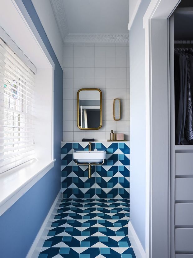 A narrow, bright bathroom viewed from the doorway, featuring a wall-mounted white basin with exposed brass plumbing, a large rounded-rectangle brass-framed mirror, a smaller matching mirror, pale blue painted walls, white tiled upper walls, and bold geometric blue, white, and dark floor and vanity-wall tiles. A window with white blinds is on the left, and an open walk-in wardrobe with hanging clothes is visible through the doorway on the right. Decorative ceiling cornices and white trims add a traditional architectural detail.