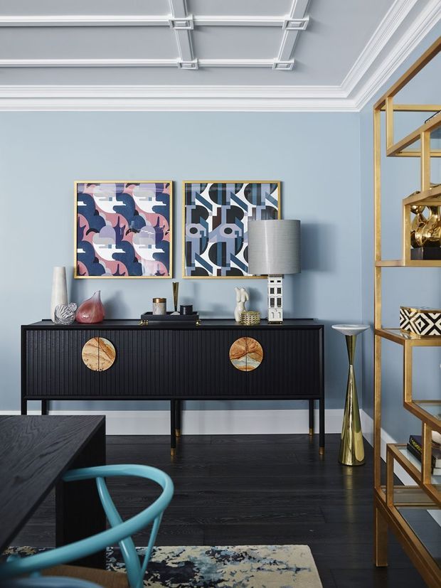 A refined, eclectic living room vignette with a black fluted sideboard, sculptural gold-accented shelving, colourful abstract artwork, decorative objects, and a dark timber floor. Pale blue walls and an ornate white coffered ceiling create a sophisticated backdrop, while a teal chair and patterned rug add bold colour and texture.