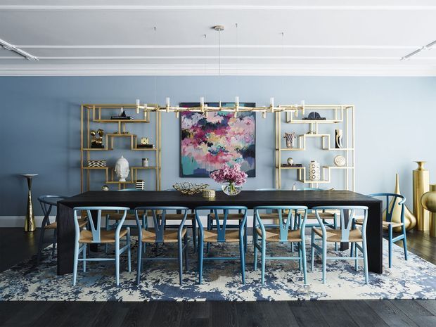 A sophisticated dining room with a long dark rectangular dining table surrounded by blue-toned chairs. The room features pale blue walls, dark timber flooring, a large patterned area rug, sculptural gold and white open shelving, and a contemporary multi-light chandelier. Abstract artwork, decorative objects, and floral arrangements add colour and visual interest.