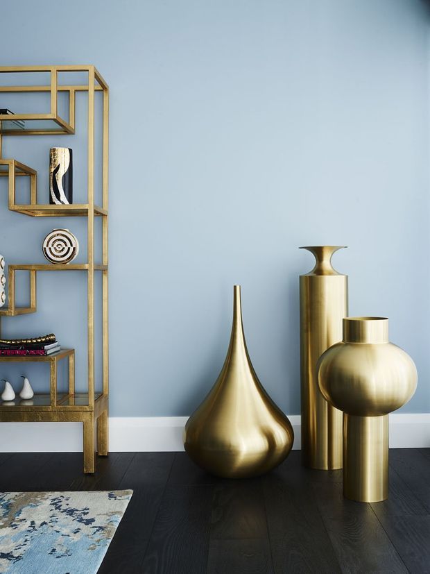 A styled interior vignette with a pale blue wall, dark timber flooring, and a tall open brass-toned shelving unit displaying decorative objects. Several large sculptural brass vases stand on the floor, with a partial blue patterned rug in the foreground. The composition is refined and decorative, with strong metallic accents and a cool blue backdrop.