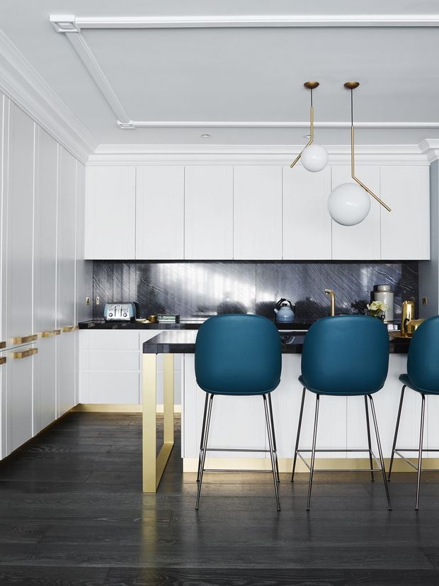 A refined contemporary kitchen with white floor-to-ceiling cabinetry, detailed white wall panelling and moulded ceiling trim. A dark stone or marble-look splashback and black countertop contrast with the deep blue upholstered bar stools. Brass or gold-toned hardware, tapware and island supports add warmth, while sculptural globe pendant lights hang above the island. The room has a polished, high-contrast and luxurious character.