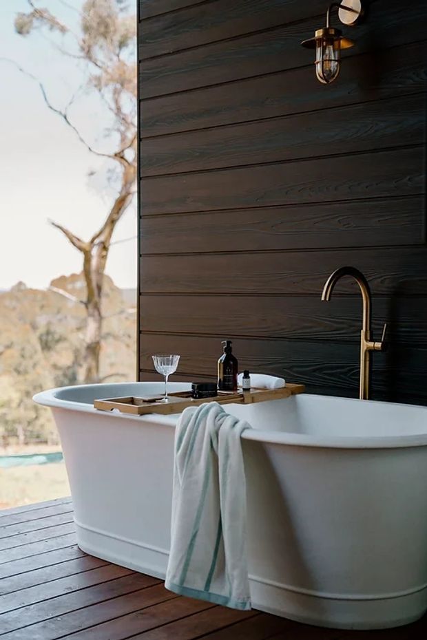 A freestanding white bathtub sits on a dark timber deck beside a dark horizontal timber-clad wall. A brass-toned curved tap, wall-mounted caged light, wooden bath tray, toiletries and folded towel create a refined outdoor bathing setting, with a dry natural landscape visible beyond the opening.
