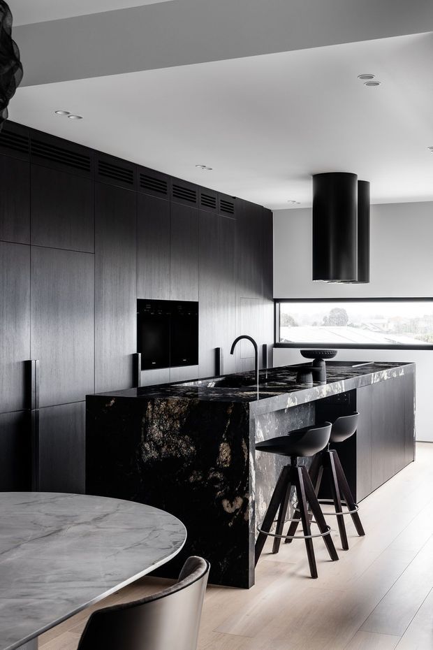 A sophisticated modern kitchen with a large dark timber wall of integrated cabinetry, black stone waterfall island and matching dark stone benchtop. Black bar stools sit at the island beneath a sculptural cylindrical black rangehood. A marble-look dining table is partially visible in the foreground, with pale timber flooring, white ceilings, recessed downlights and broad horizontal glazing.