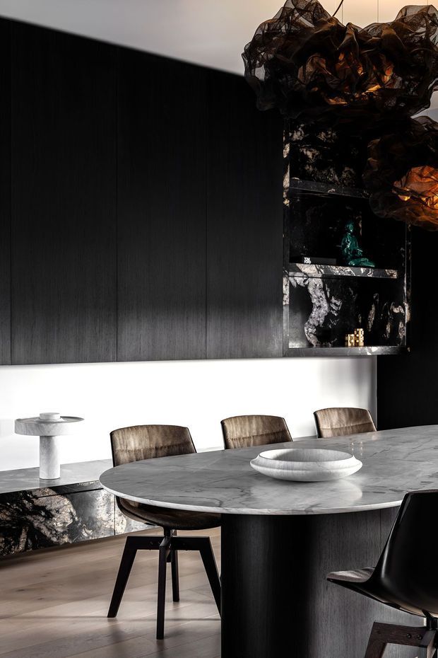 A sophisticated contemporary dining area featuring a large round stone dining table with dark sculptural base, upholstered dining chairs, dark vertically paneled cabinetry, and dramatic black marble shelving and wall elements. Sculptural pendant lighting and restrained decorative objects create a moody, luxurious atmosphere.