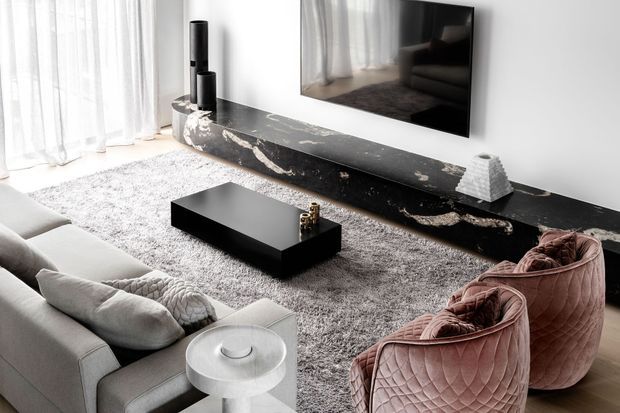 A refined contemporary living room with a large white sectional sofa, two blush-pink quilted lounge chairs, a low black rectangular coffee table, a textured grey area rug, and a dramatic black marble-look linear media console with integrated wall-mounted television. Sheer curtains provide soft natural light, while black decorative objects and a sculptural white vase add contrast.