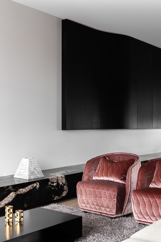 A refined contemporary living room with a restrained monochrome palette and luxurious accents. Matte black, vertically fluted built-in cabinetry spans the upper wall, while a long dark stone console runs beneath it. Two blush-pink quilted lounge chairs sit on a deep grey shag rug around a low black coffee table. Decorative objects include a faceted white sculpture and small gold-toned candleholders. The room has a clean, sculptural and moody character.