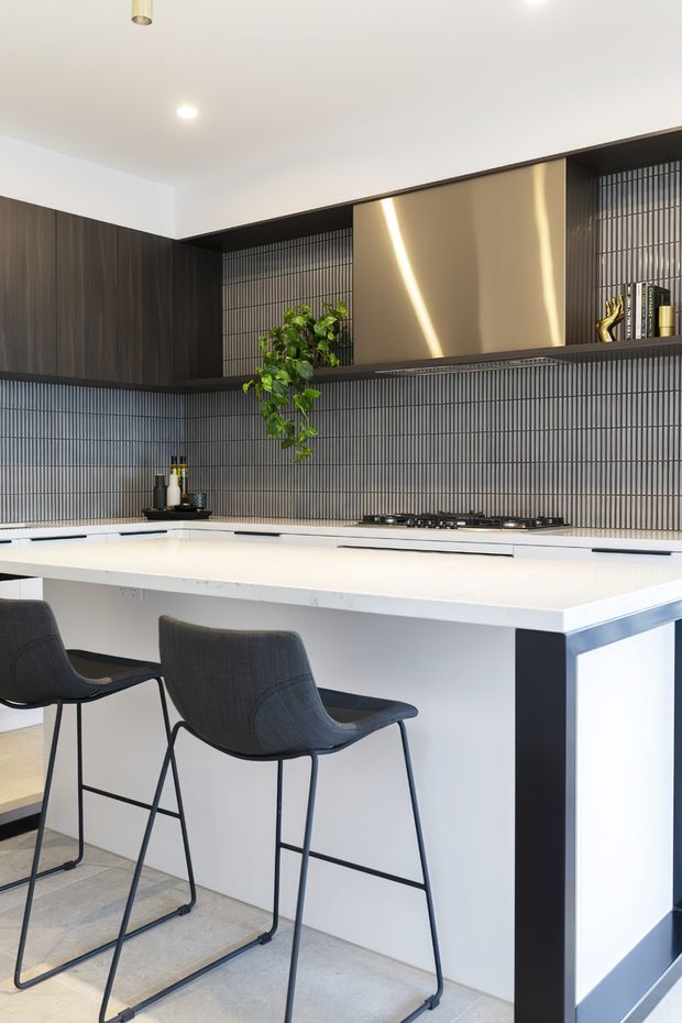 Contemporary kitchen featuring a large white island with integrated seating, dark timber cabinetry, a vertically ribbed dark backsplash, open shelving, and warm metallic feature panels. Black bar stools and subtle greenery add contrast to the bright, refined space.