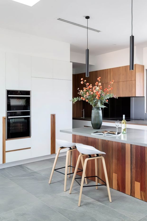 Contemporary kitchen interior with a large island and two sculptural bar stools. The space features full-height white cabinetry with integrated black ovens, rich vertical-grain timber cabinetry, a pale grey stone island benchtop, dark pendant lights, and large grey stone floor tiles. A sink, open cookbook, wine bottle and vase of flowers sit on the island, while a dark splashback and television or reflective appliance are visible at the rear.