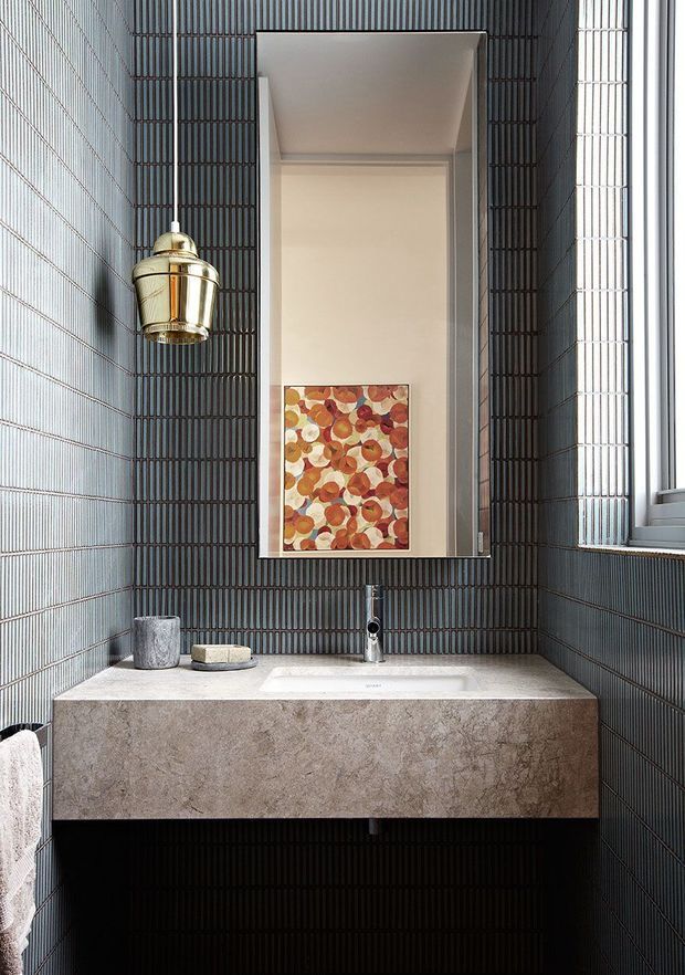 A compact contemporary bathroom featuring dark vertically ribbed wall tiles, a wall-mounted stone vanity with an integrated white basin, a slim chrome tap, a large rectangular mirror, a brass wall light, and a small abstract artwork reflected in the mirror. Natural light enters through the window at right.