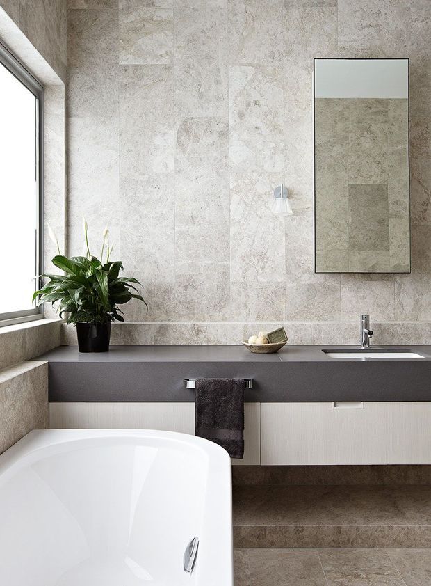 A refined contemporary bathroom featuring large-format beige-grey stone-look wall and floor tiles, a freestanding white bathtub, wall-mounted dark grey vanity with pale textured cabinetry, an integrated basin, frameless rectangular mirror, and a potted plant beside a bright window.