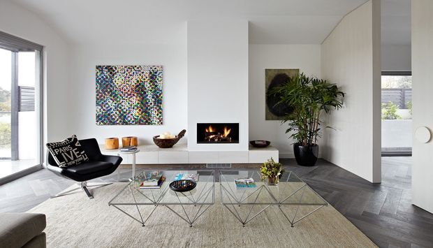 Bright contemporary living room with a sleek black lounge chair, glass geometric coffee tables, a large neutral rug, wall-mounted fireplace, colourful artwork, indoor plants, and extensive glazing. The room has a clean white palette with dark herringbone flooring and views to the outdoors.