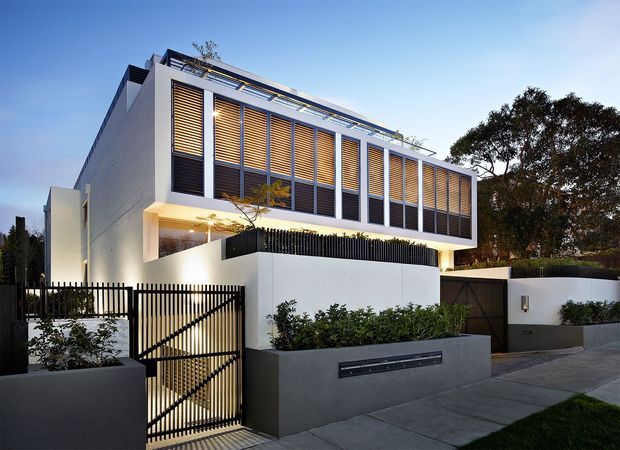 Contemporary two-storey residential facade with crisp white rendered walls, a projecting upper-level volume, extensive dark-framed glazing screened by horizontal timber louvres, and a sheltered illuminated entry behind a black vertical-slat pedestrian gate. A dark grey rendered boundary wall, integrated letterbox, landscaped planting, driveway paving, and mature trees complete the street frontage.