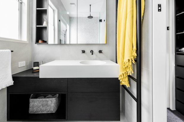 A contemporary bathroom vanity with a white integrated basin and wall-mounted black fixtures, set against grey textured walls. The vanity features dark timber cabinetry with open storage, a large mirrored cabinet, and a glass-enclosed shower visible behind it. A bright yellow towel adds a bold accent beside the black-framed shower enclosure.