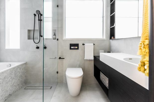 Bright contemporary bathroom with a frameless glass walk-in shower, wall-hung toilet, freestanding-style white bathtub, and dark floating vanity. Light grey tiled surfaces, recessed shower shelving, and a vivid yellow towel provide contrast.