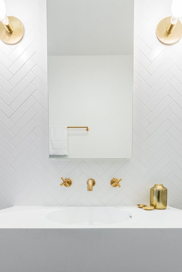 A bright, refined bathroom vanity featuring a large frameless mirror, white herringbone wall tiles, a white integrated basin and countertop, and brushed gold wall-mounted tapware and sconces. A grey floating vanity cabinet and small gold decorative accessories add contrast.