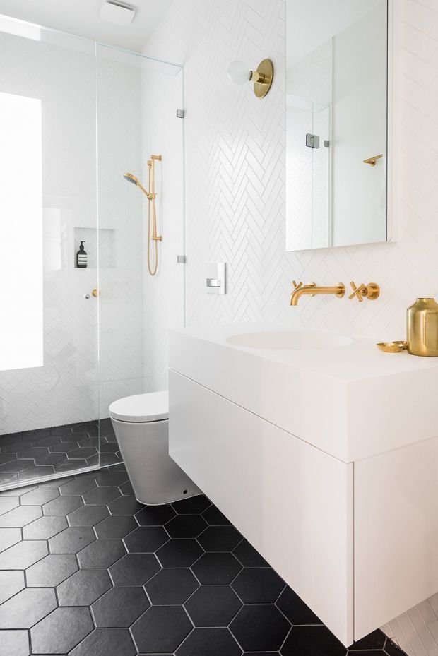 Bright contemporary bathroom featuring a frameless glass walk-in shower, white herringbone wall tiles, dark charcoal hexagonal floor tiles, a floating white vanity with integrated basin, mirrored wall cabinet, wall-mounted gold fixtures, and a white toilet. The palette is predominantly white and black with warm brass-gold accents.