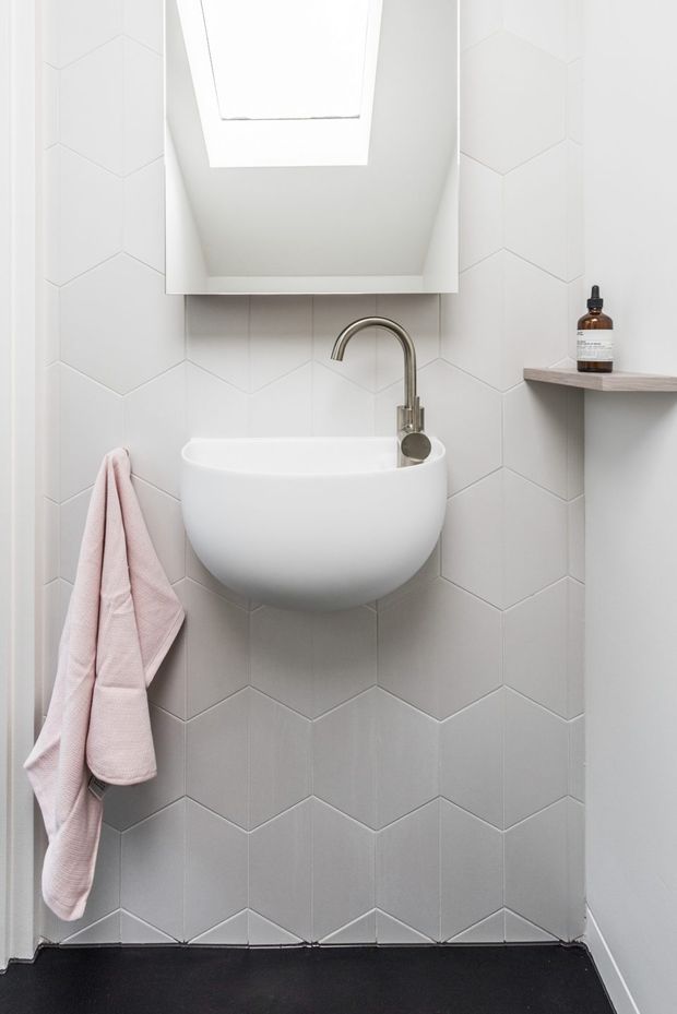 Compact, bright bathroom featuring a wall-mounted rounded white basin with a brushed metal gooseneck tap, pale geometric chevron wall tiles, a skylight, a small floating shelf with soap, and a soft pink towel. The floor is a dark matte finish, creating contrast with the predominantly white and light grey surfaces.