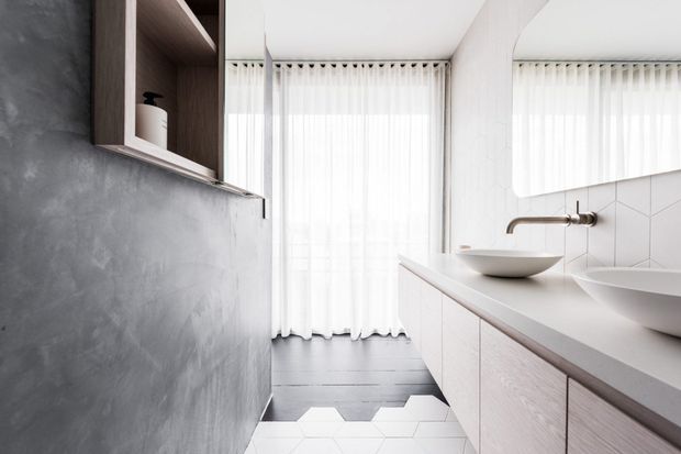Contemporary bathroom with a long double vanity, two vessel basins, wall-mounted brushed metal tapware, pale cabinetry, a large frameless mirror, and white geometric wall and floor tiles. A textured grey plaster wall and full-height sheer curtains add contrast, while open timber shelving provides storage.