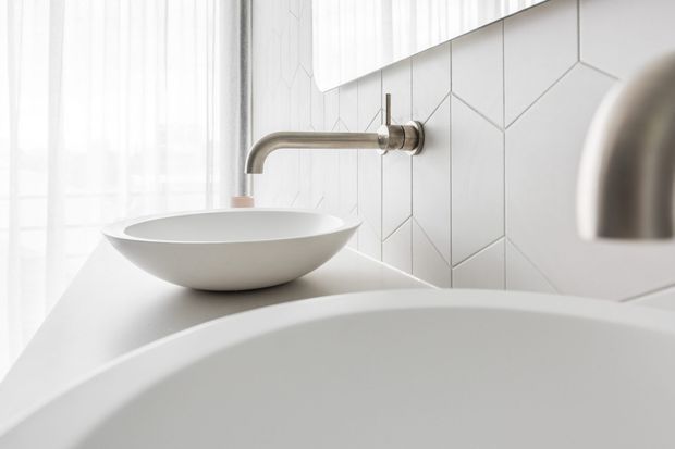 Bright minimalist bathroom detail featuring a white vessel basin on a pale vanity, a wall-mounted brushed metal tap, geometric white wall tiles, sheer floor-to-ceiling curtains, and the curved edge of a freestanding bathtub in the foreground.