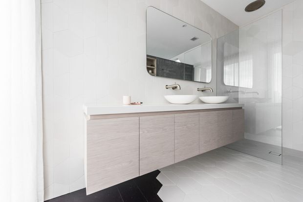 Bright contemporary bathroom featuring a floating double vanity with pale timber-look cabinetry, white vessel basins, wall-mounted brushed metal tapware, a large mirror, white geometric wall tiles, and a frameless glass shower enclosure. Light grey floor tiles and a dark contrasting floor section create subtle visual contrast.