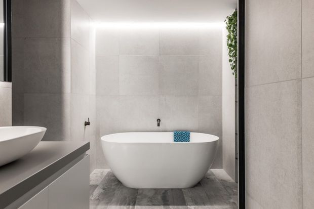 Contemporary bathroom featuring a freestanding white oval bathtub, large light-grey wall tiles, grey stone-look flooring, a minimalist dark vanity edge, concealed warm LED cove lighting, and a narrow vertical planter detail.
