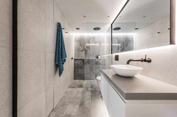 Contemporary bathroom with a walk-in shower enclosed by glass, large-format grey stone-look wall and floor tiles, a floating white vanity with a broad grey benchtop, vessel basin, wall-mounted bronze tapware, and a large illuminated mirror. Recessed ceiling lighting and concealed LED lighting create a clean, refined atmosphere.