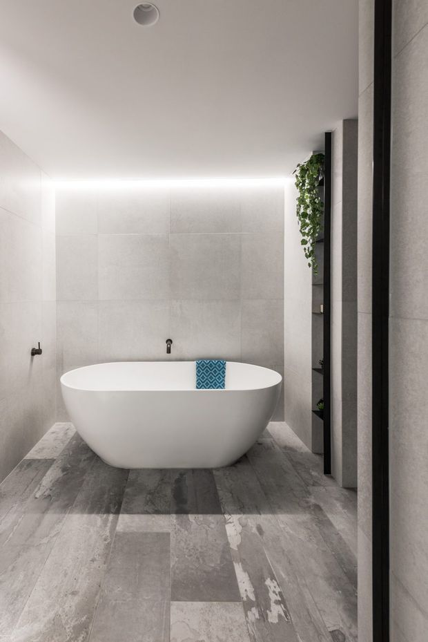 Contemporary minimalist bathroom featuring a freestanding white oval bathtub, large-format pale grey wall tiles, grey stone-look floor tiles, recessed ceiling lighting, and a dark open shelving unit with trailing greenery.