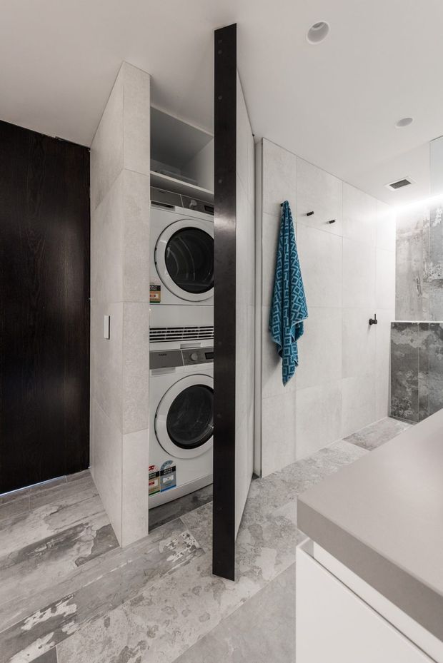 Compact contemporary bathroom with an integrated concealed laundry cupboard containing stacked front-loading washer and dryer. The room features large pale grey stone-look wall and floor tiles, white built-in vanity cabinetry with a grey stone benchtop, dark timber door and vertical dark joinery detail, recessed ceiling lights, and a walk-in shower area with wall-mounted towel hooks.