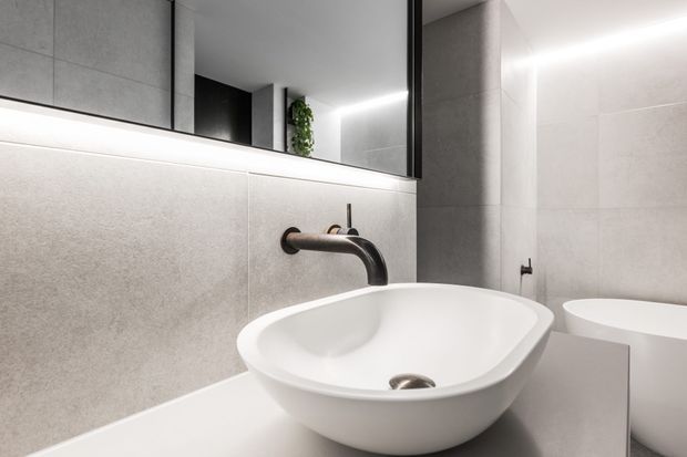 Contemporary bathroom vanity featuring a white vessel basin on a pale stone-look benchtop, wall-mounted brushed dark-metal tapware, large-format light grey wall tiles, and a wide mirrored cabinet with integrated linear lighting. A freestanding bathtub is partially visible in the background.