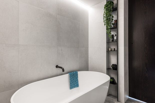 Contemporary bathroom featuring a freestanding white bathtub against large-format light grey wall tiles. A matte black wall-mounted tap, recessed open shelving with toiletries and trailing greenery, and a dark timber-toned door create a refined, modern scheme. Soft concealed lighting adds a calm atmosphere.