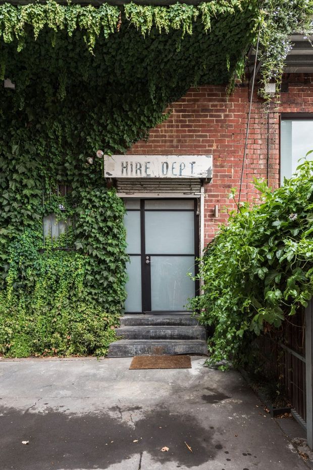 A leafy urban commercial entrance framed by dense climbing ivy and shrubs. The facade combines exposed red brick with a dark-framed frosted-glass door, concrete steps, and a weathered sign reading “MIRE DEPT,” creating a tucked-away, warehouse-like character.