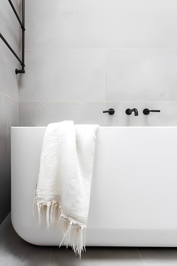 A minimalist bathroom featuring a freestanding white bathtub with a draped white fringed towel, large pale grey wall tiles, matte black wall-mounted bath tapware, and a simple black wall rail. The space has a clean, monochromatic contemporary appearance.