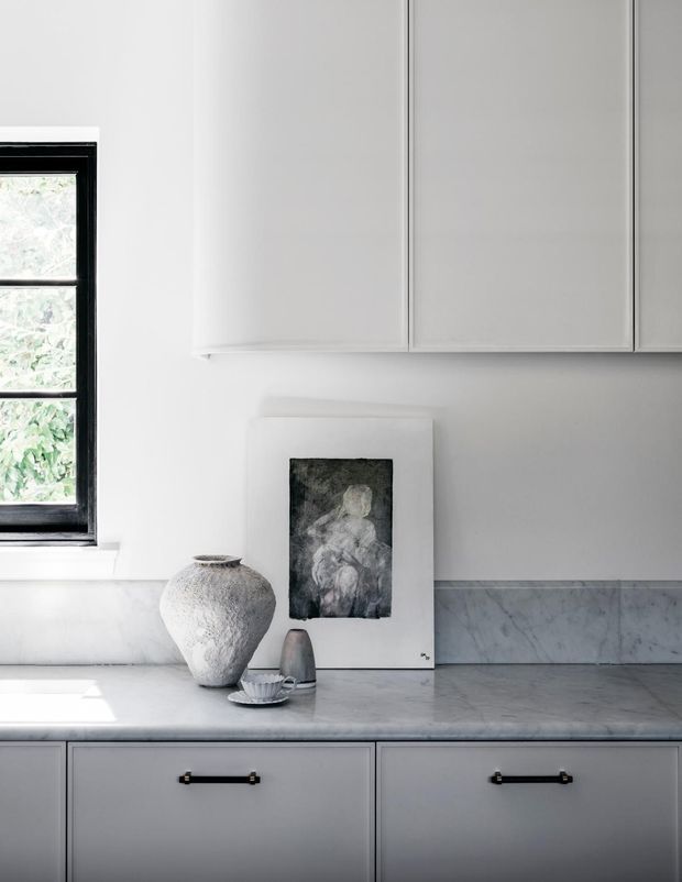 A close-up view of a refined, monochromatic kitchen featuring pale grey-white cabinetry, a grey veined stone benchtop and splashback, black-framed window, and dark metal cabinet handles. Neutral sculptural decor and framed artwork rest on the benchtop, creating a calm, minimalist composition.