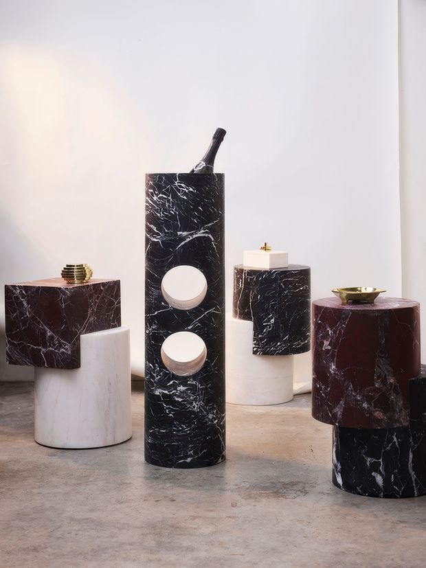 A curated product-style display of sculptural cylindrical and rectangular forms in richly veined black, burgundy, and white marble or stone, arranged on a concrete floor against a plain white wall. Small metallic decorative objects and a bottle add accents, creating a refined, gallery-like composition.