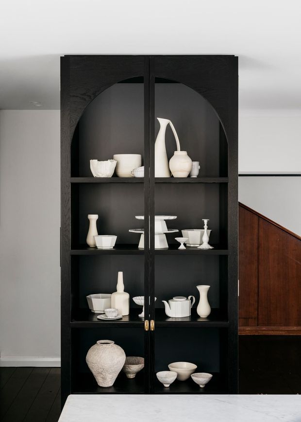 A tall, black built-in display cabinet with arched internal openings and glass doors, filled with curated white ceramic and stoneware vessels. The cabinet stands against a pale wall beside a dark timber staircase, with a dark timber floor and a light stone surface in the foreground. The composition is refined and minimalist, with strong black-and-white contrast and warm timber accents.