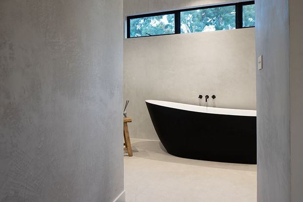 Minimalist bathroom viewed through a doorway, featuring a sculptural freestanding black-and-white bathtub, wall-mounted dark fixtures, pale textured plaster walls, light flooring, and a narrow horizontal window overlooking trees.