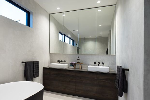 Contemporary bathroom featuring a long dark timber double vanity with two white vessel basins, wall-mounted black fixtures, a large mirrored cabinet, textured light plaster walls, recessed ceiling lights, and a freestanding white bathtub partially visible in the foreground. High horizontal windows provide natural light.