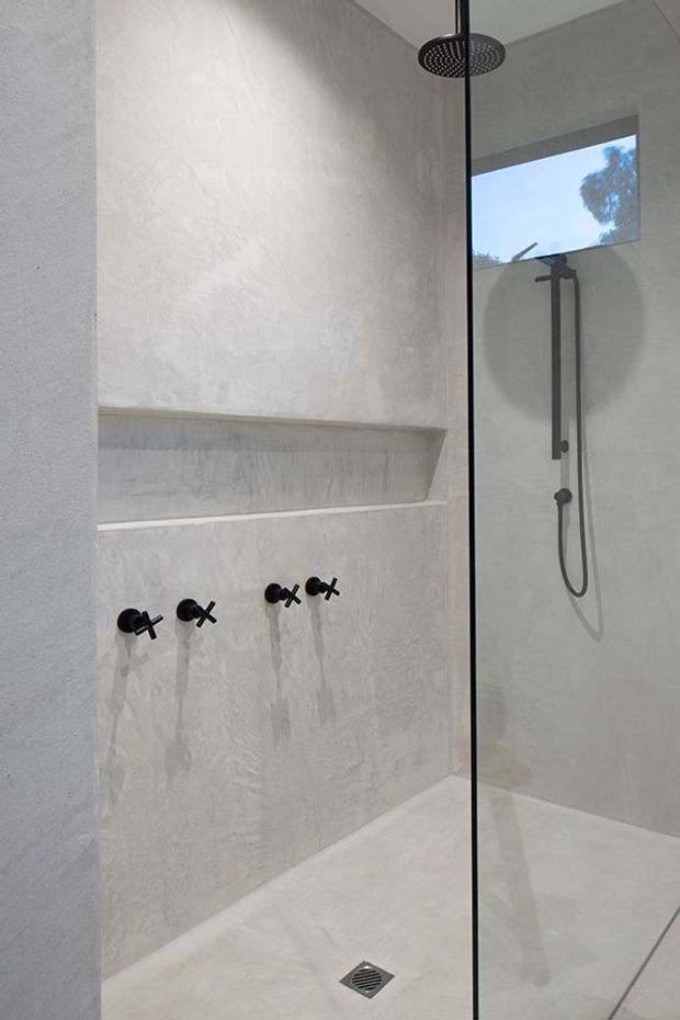 Minimalist walk-in shower with a frameless glass screen, textured light plaster or microcement walls, integrated recessed shelf, matte black shower fittings, overhead rain shower, handheld shower rail, and a linear floor drain. Natural light enters through a high window.