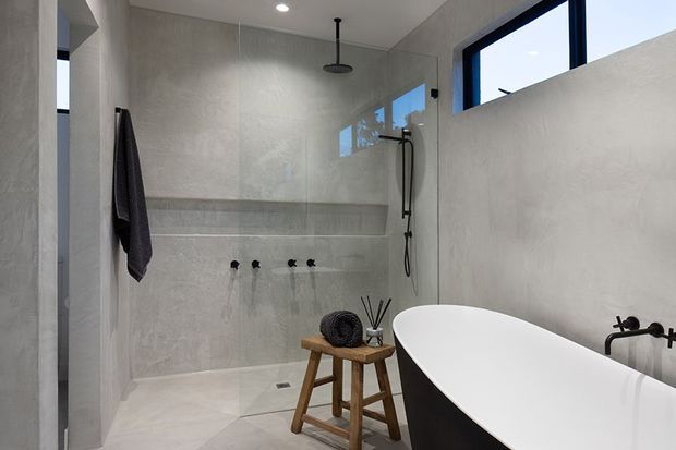 A contemporary minimalist bathroom featuring large pale grey concrete-look wall and floor finishes, a walk-in shower with recessed shelving and matte black fixtures, high-set black-framed windows, and a freestanding white bathtub with a dark exterior. A small timber stool adds warmth to the otherwise restrained palette.